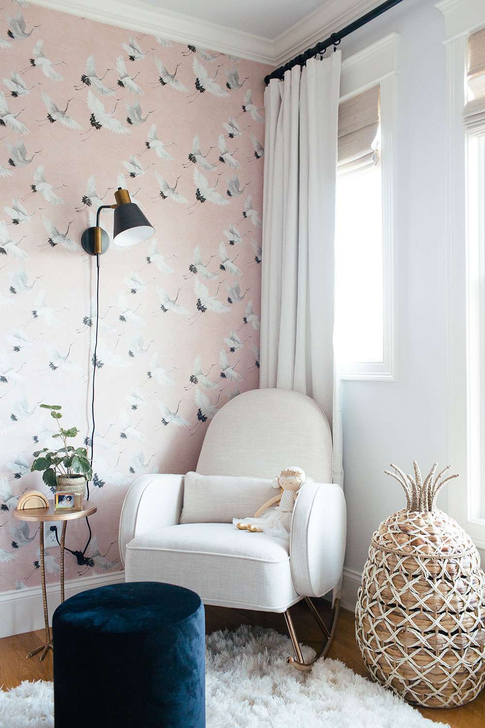 nursery with pink crane wallpaper accent wall, black sconce, white rocking chair with wicker pineapple basket