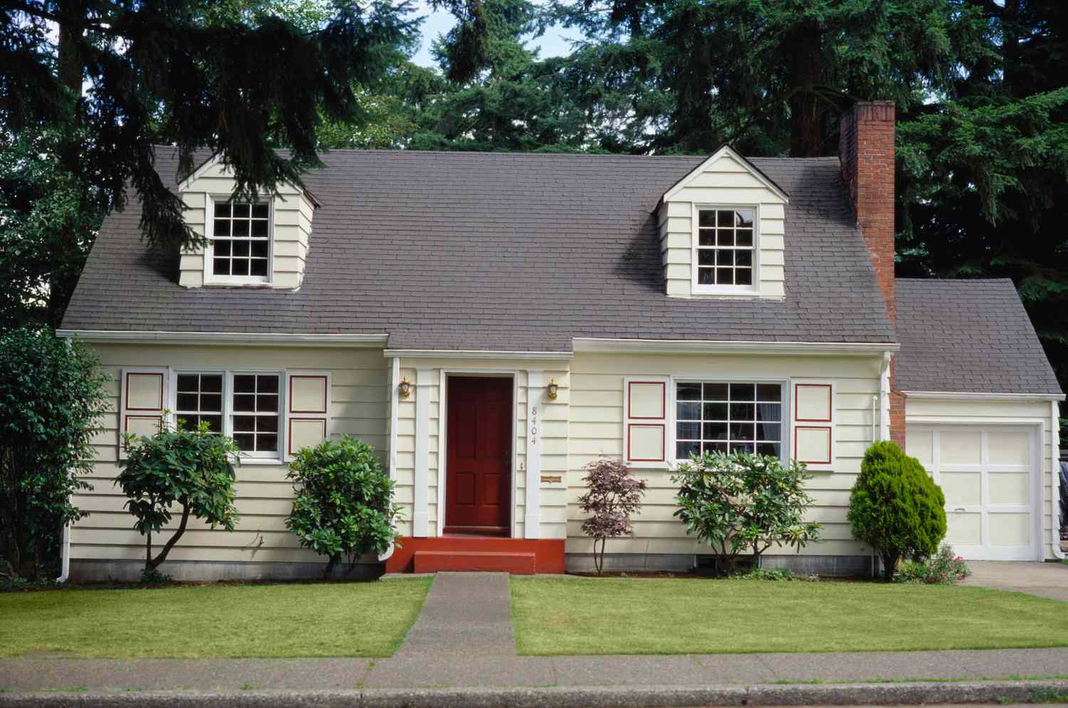 Fassade eines Hauses im Cape-Cod-Stil mit zwei Dachgauben, seitlichem Kamin und Garage mit 1 Erker, mit mehrverglasten Fenstern und geometrisch gemusterten Fensterläden
