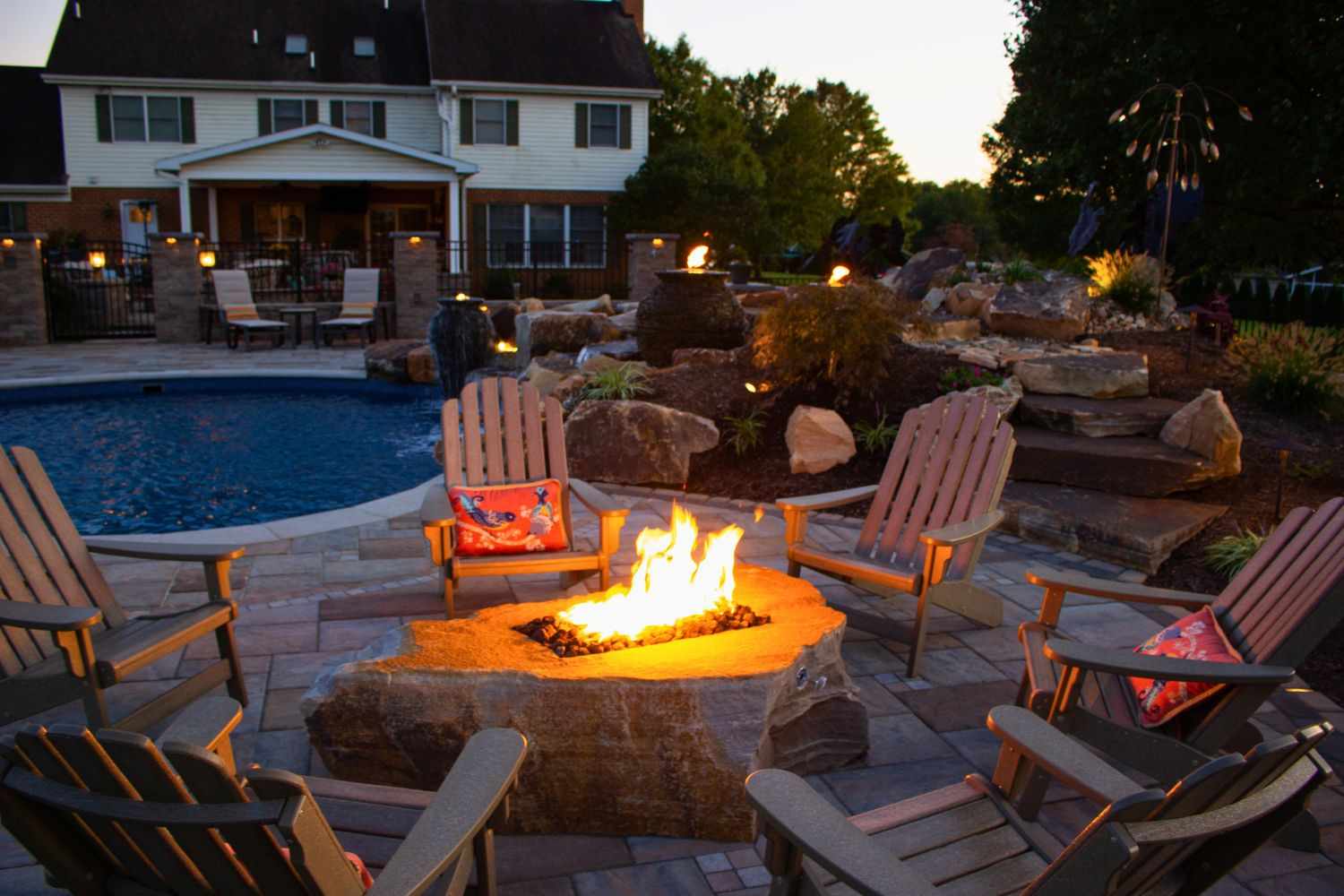 Wie man in diesem Sommer eine DIY -Feuerstelle entwirft, die tatsächlich funktioniert 16 Eine von Holzstühlen in einem Garten mit einem Pool umgebene natürliche förmige Steingrube umgeben