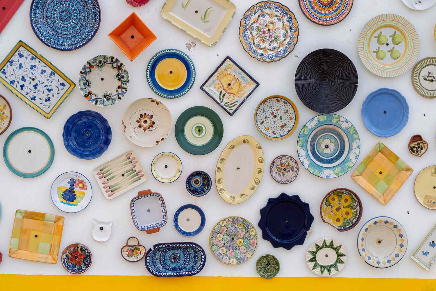 Sammlung von farbenfrohen Keramikplatten und -gerichten, die in Formen, Größen und Designs variieren, werden an einer weißen Wand angezeigt, die eine lebendige und künstlerische Anordnung zeigt